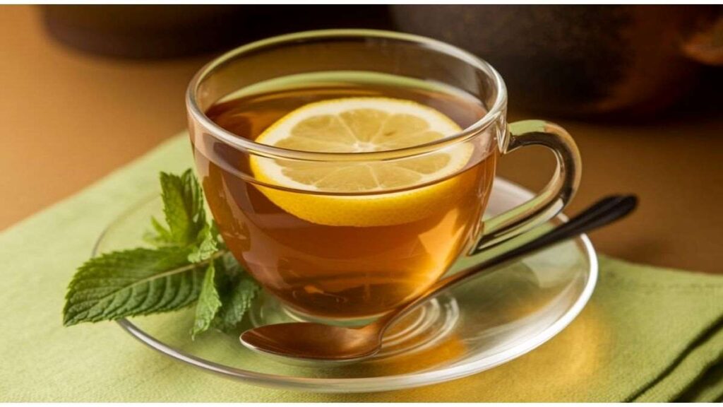 Glass cup of lemon tea with a lemon slice, mint leaves, and a spoon on a glass saucer, placed on a green napkin.