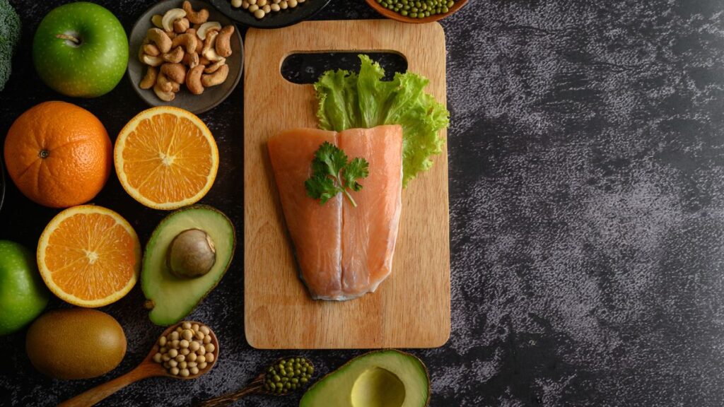 A wooden cutting board with fresh salmon and lettuce, surrounded by healthy foods like oranges, avocados, nuts, and soybeans, ideal for an acne free diet.